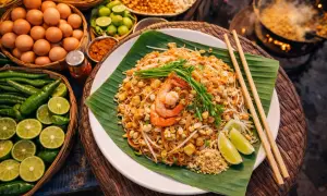 Talerz Pad Thai z krewetkami, posypany orzeszkami, na liściu bananowca, obok składniki.