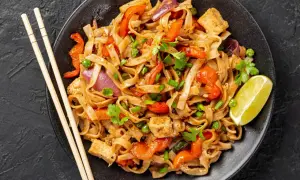 Talerz pad thai z makaronem ryżowym, warzywami, tofu i limonką, obok pałeczki.