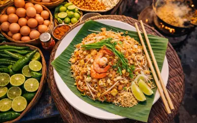 Talerz Pad Thai z krewetkami, posypany orzeszkami, na liściu bananowca, obok składniki.