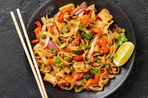 Talerz pad thai z makaronem ryżowym, warzywami, tofu i limonką, obok pałeczki.
